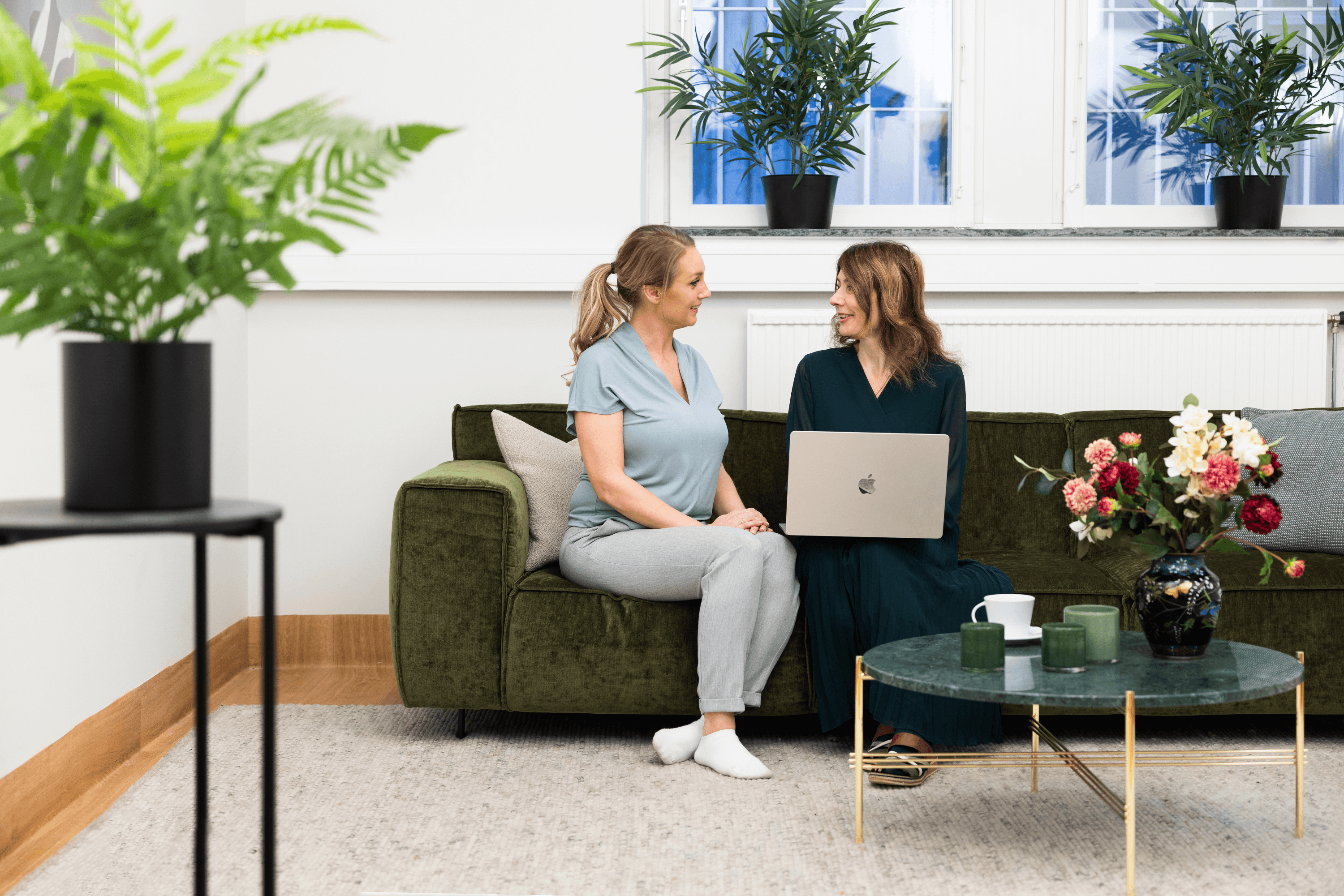 Närbild på ett bord i ett patientrum med en vattenkanna, ett glas vatten och en liten blomma i en brun glasvas. I bakgrunden syns sängkläder med blommigt mönster.