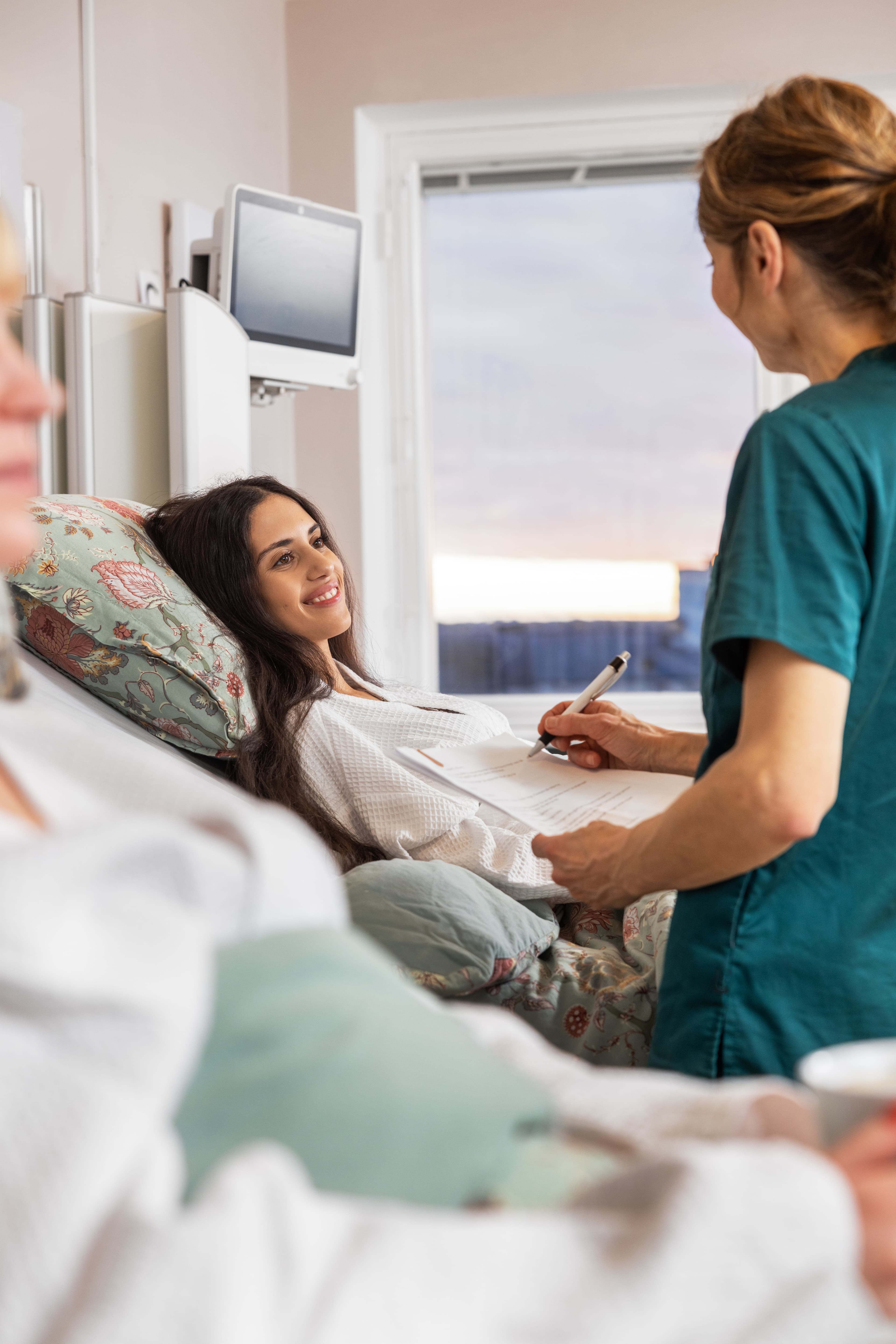 En ung kvinna med mörkt hår ligger i en sjukhussäng och ler mot en kirurg i gröna operationskläder som står bredvid och håller ett formulär. Bakgrunden visar en rosa och blå solnedgång genom ett fönster, och en annan patient är delvis synlig i förgrunden.