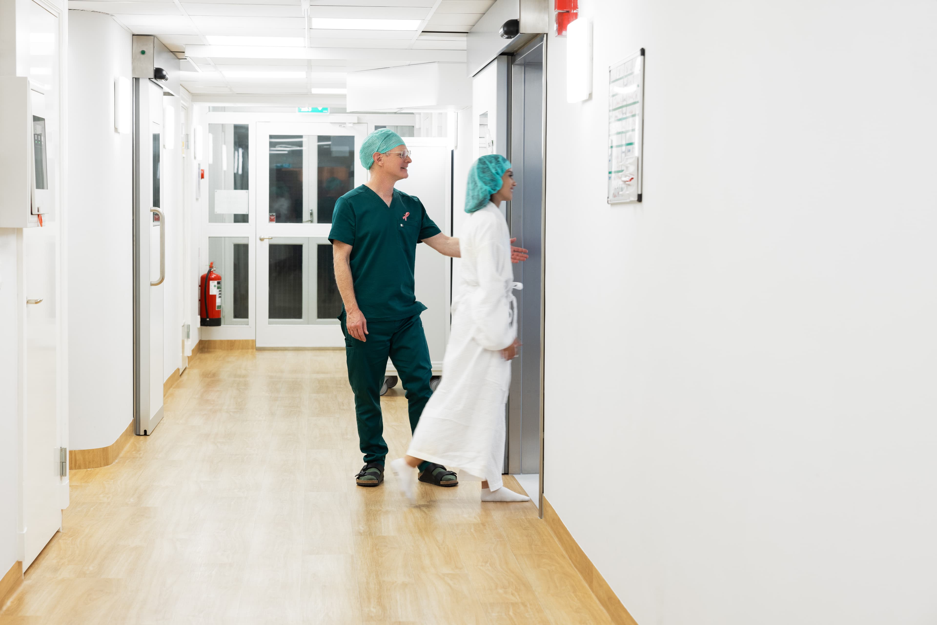 En kirurg i gröna operationskläder och grön operationsmössa följer en patient i vit morgonrock och grön operationsmössa genom en sjukhuskorridor med ljust trägolv och ljus belysning.