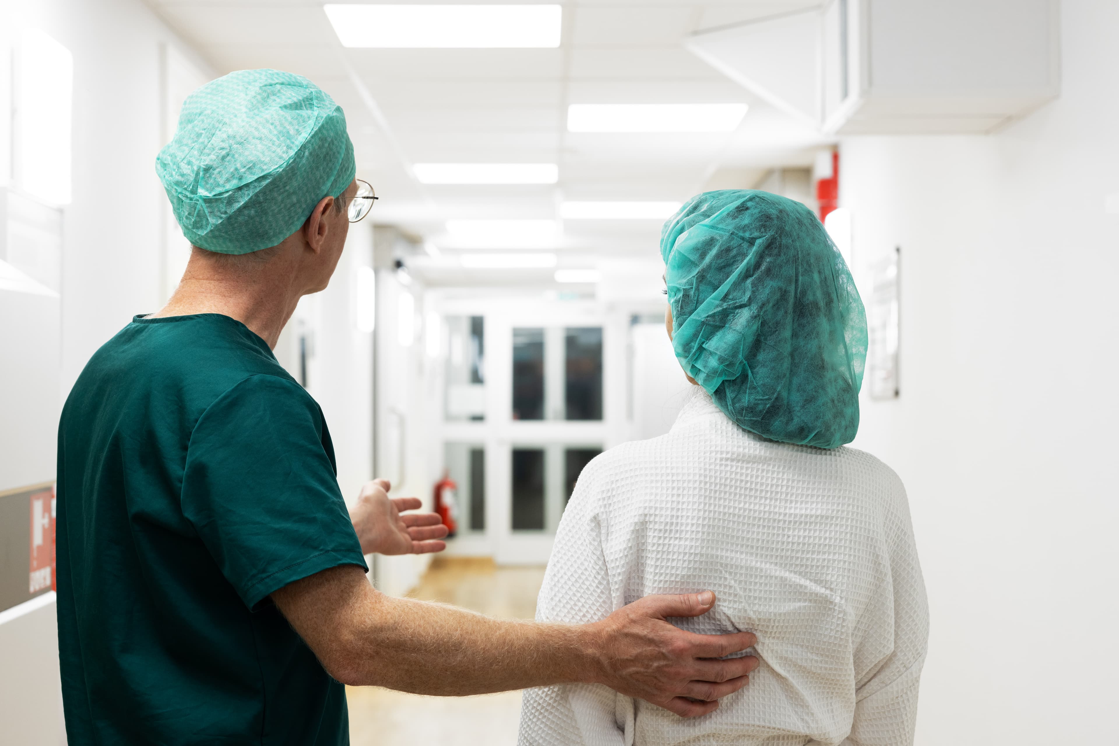 En kirurg i gröna operationskläder visar vägen för en patient klädd i vit morgonrock och grön operationsmössa i en sjukhuskorridor med ljus belysning.